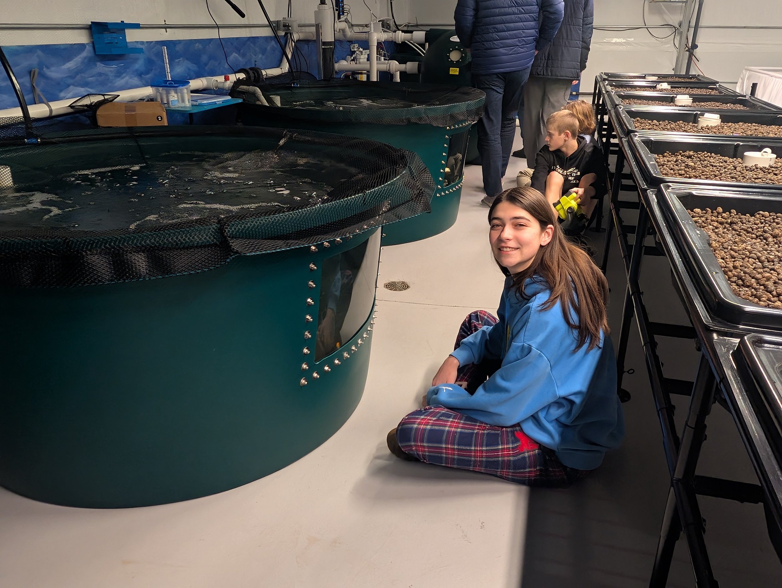 A young girl sitting on the floor in a research facility, smiling at the camera. She is wearing a blue hoodie and plaid pants, surrounded by large circular tanks filled with water and tables displaying various materials. Two other children are seated nearby, and adults are visible in the background, emphasizing a collaborative learning environment focused on aquaculture or marine biology.