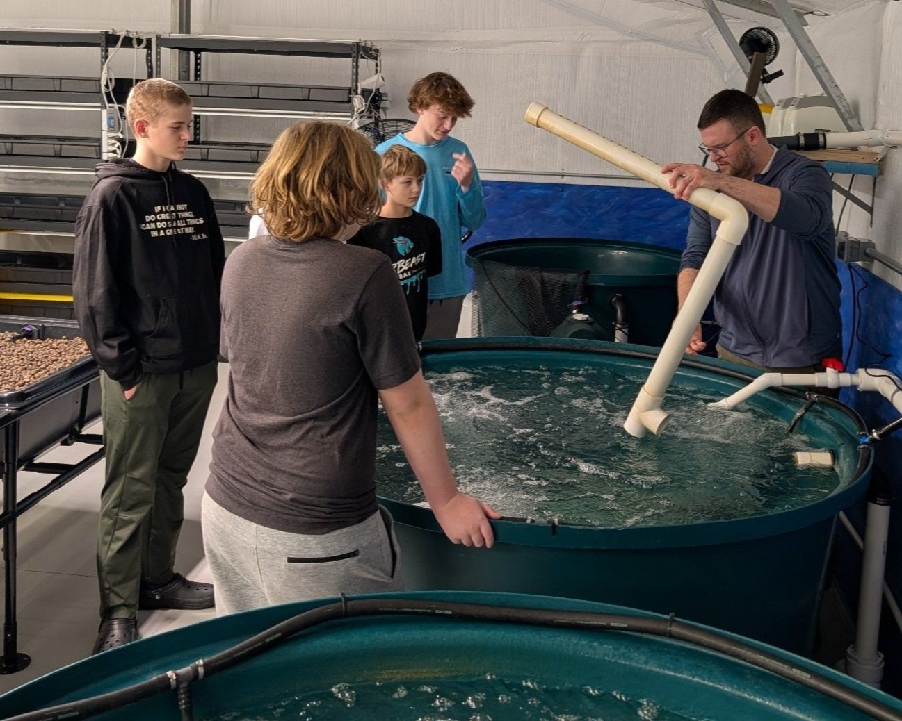 Group of young students observing a man demonstrating water flow in large aquaculture tanks during an educational workshop on sustainable fish farming practices.